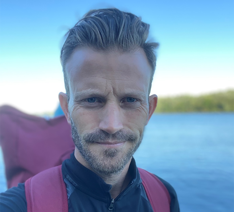 A close up Andy Caine face. He is wearing a rucksack training for his walk from John O’Groats to Land’s End. There are fields are trees blurred in the background.